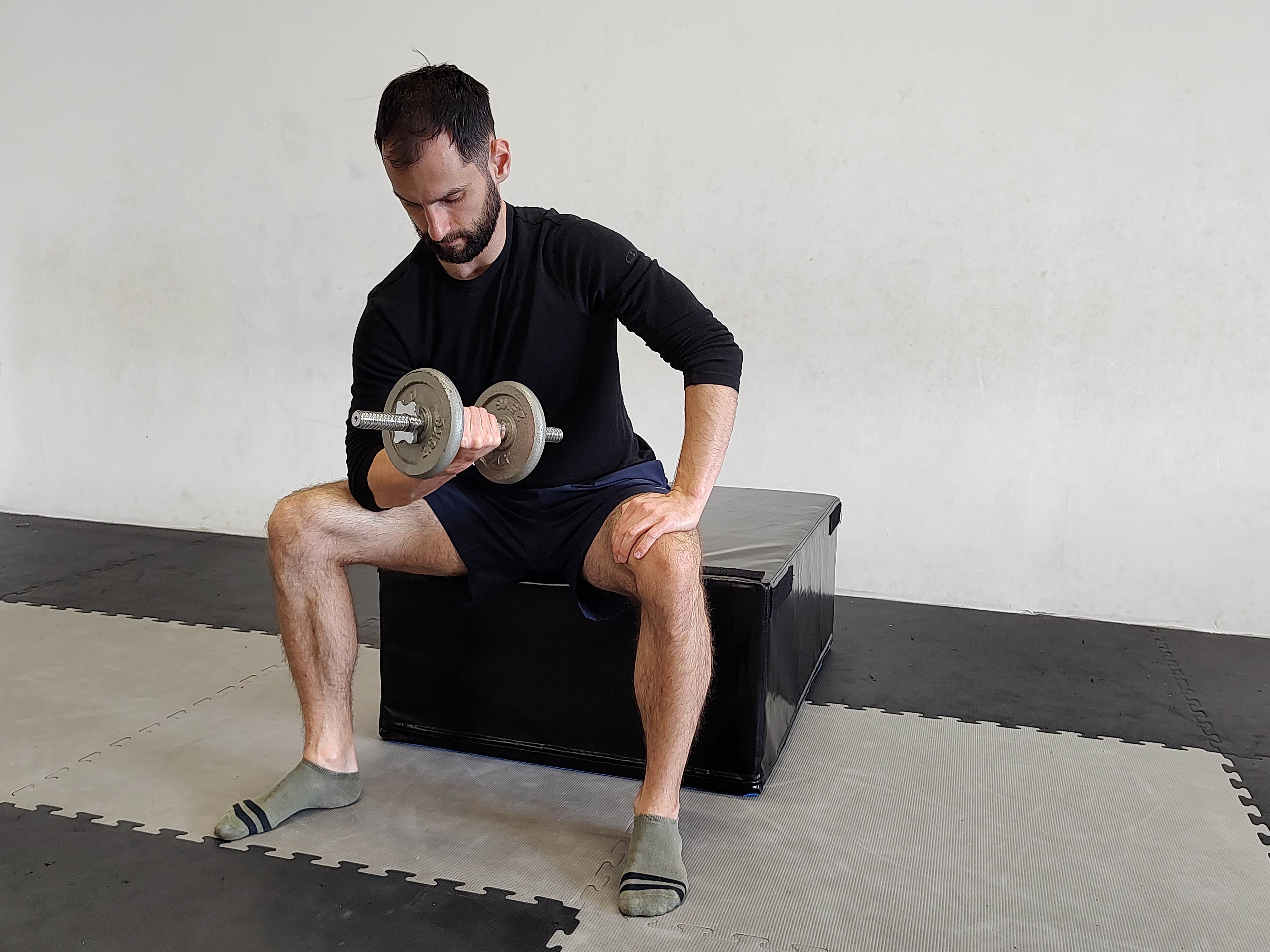 Muž v tělocvičně cvičí bicepsový zdvih s jednoručkou s oporou o koleno